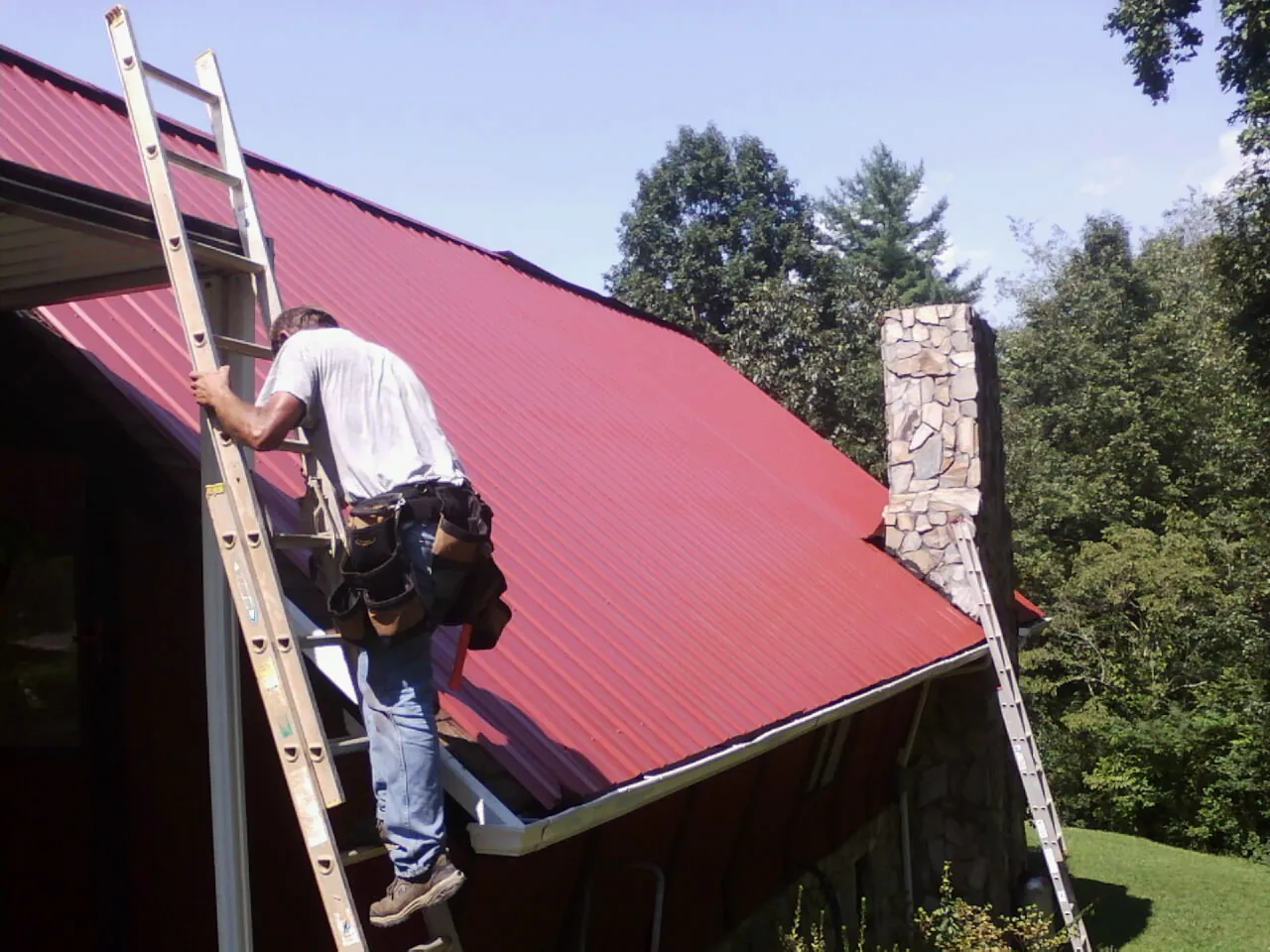Roofer on a ladder beside a red metal roof and stone chimney — Soffit & Fascia Repair by Roofing Brothers in Cowan